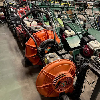A row of various lawnmowers and leaf blowers in a storage area. Prominently featured are bright orange blowers with black handles, reflecting a utilitarian tone.