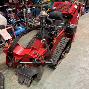 A red stump grinder with a powerful engine and rubber tracks is parked indoors. Surrounding the machine are various tools and equipment on a concrete floor.