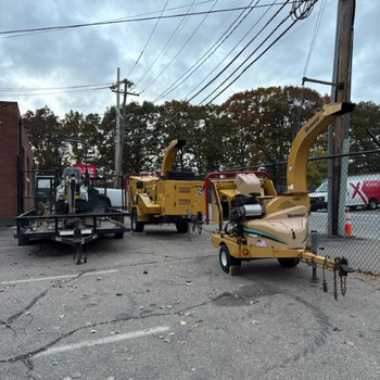 Three yellow wood chippers are parked in a fenced lot beside a brick building. Overcast sky and autumn trees in the background create a somber tone.