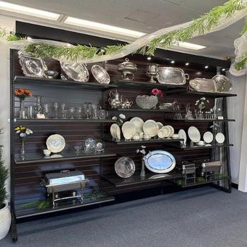 A display of elegant silver and glassware items on shelves, including platters and goblets. Floral accents and garlands add a festive touch.