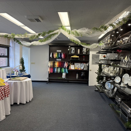 Party & Event RentalsA showroom with hanging decorations, showcasing colorful napkins, plates, and glasses on shelves. Tables display white and checkered linens. Bright, organized space.