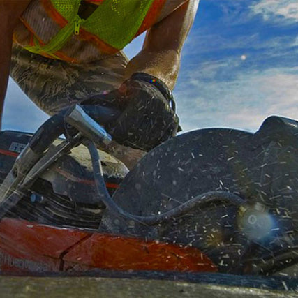 Contractor RentalsA construction worker in a safety vest and gloves operates a circular saw, cutting through concrete. The image captures dynamic motion and flying debris.